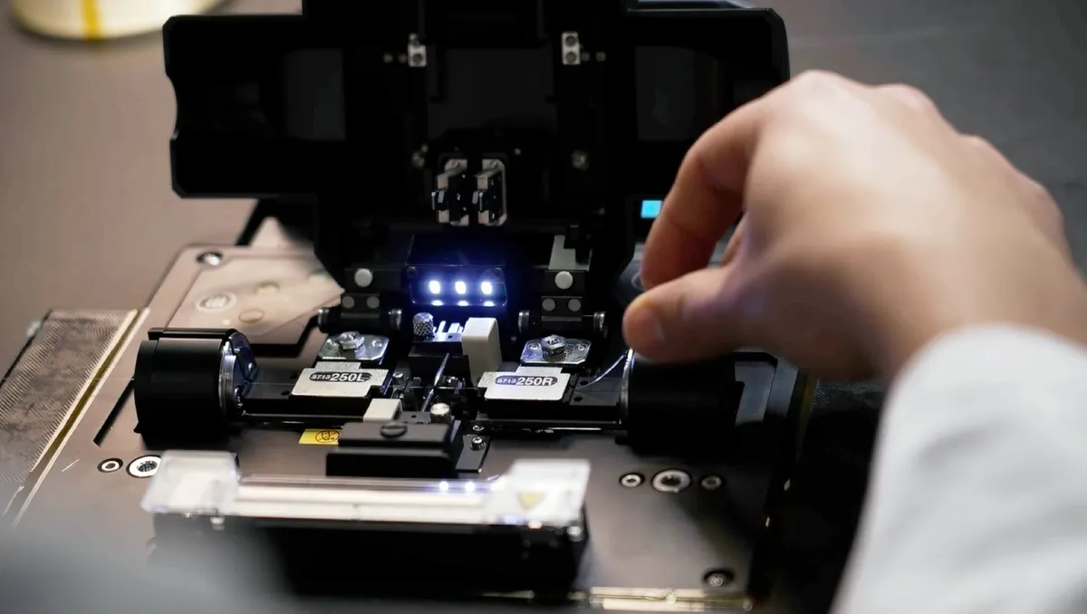 A technician's hand operates a fiber optic fusion splicer with illuminated internal components and clamps.
